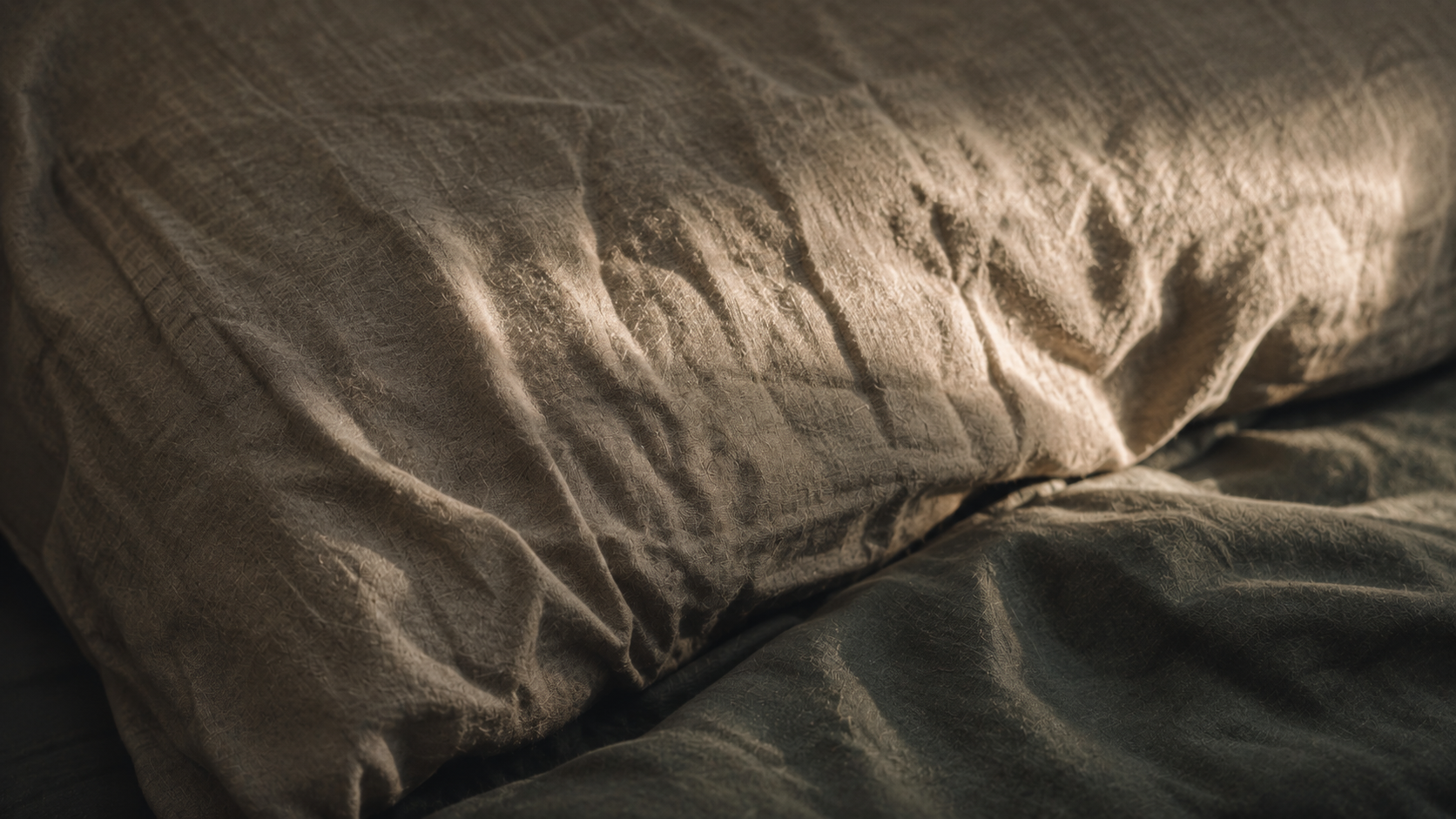Crumpled linen pillowcase in low raking morning light showing deep crease lines across the fabric