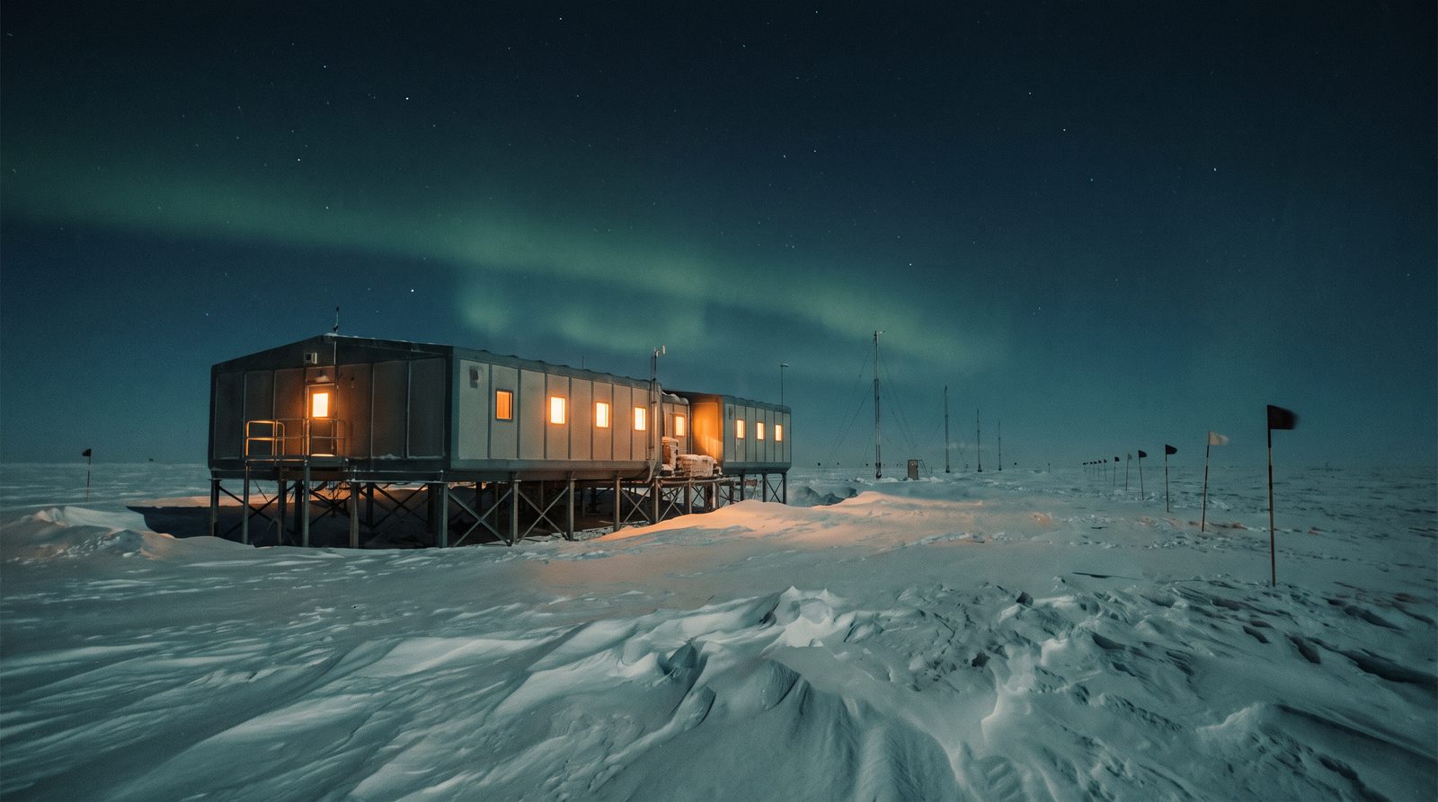 A small Antarctic research station on stilts over snow, windows glowing amber under a dark sky with faint aurora