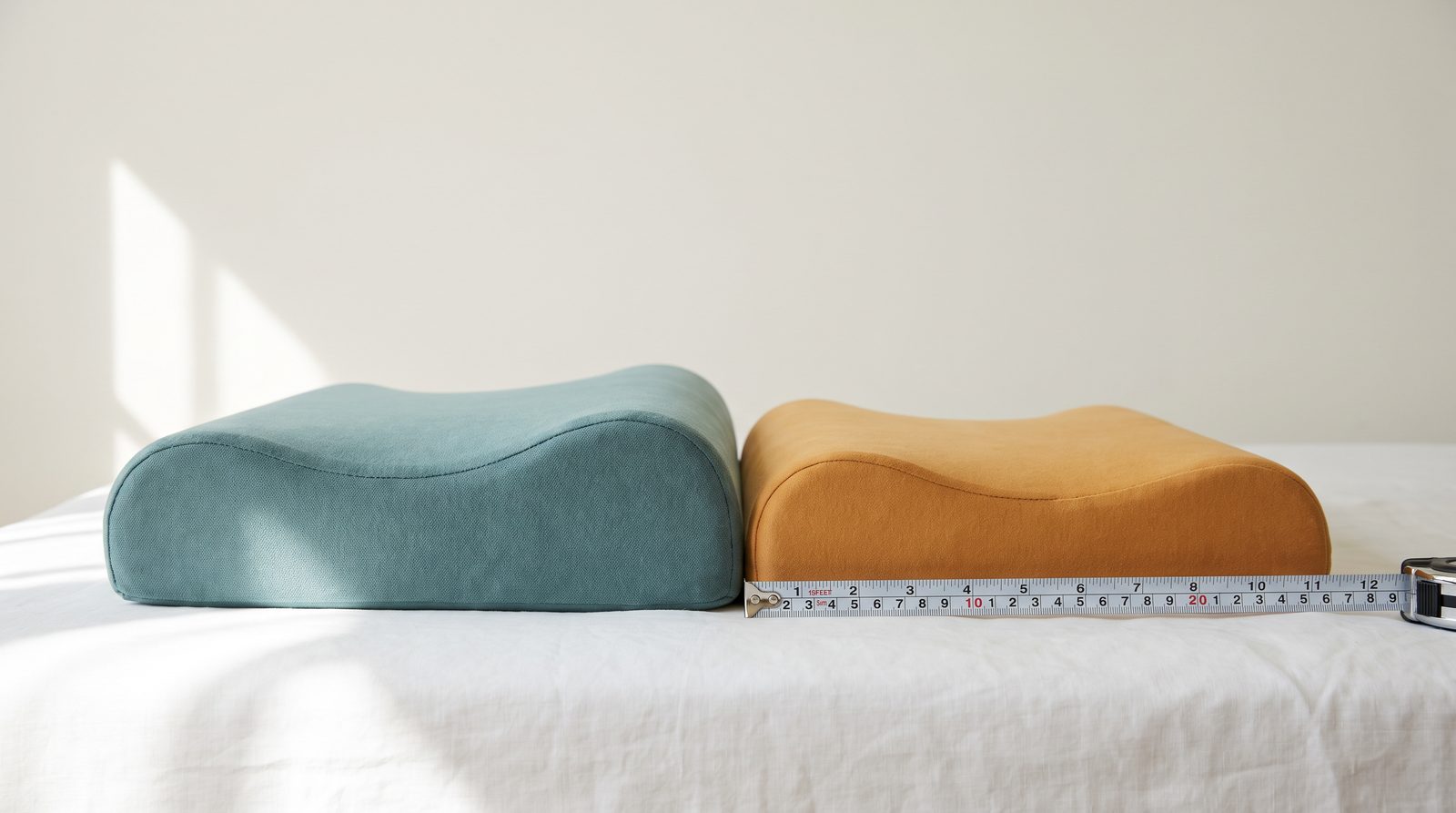 Two contoured memory foam pillows side by side on a white sheet, one clearly taller, with a tape measure beside them