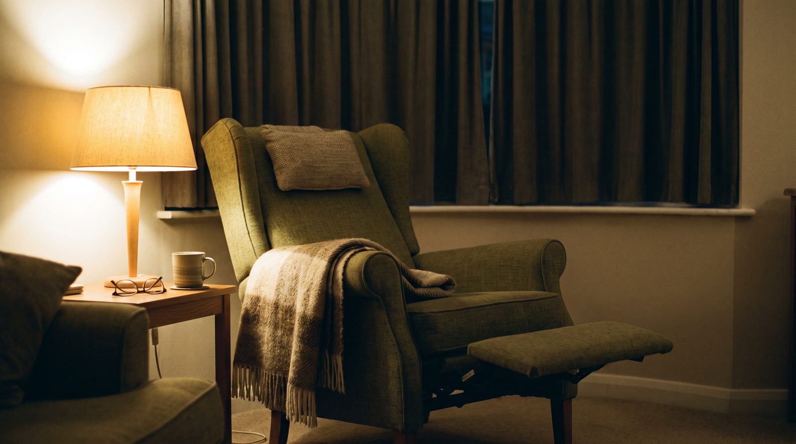 A high-backed armchair reclined with its footrest raised, a throw blanket and a small neck cushion at the headrest, lit by a single lamp in a darkened living room