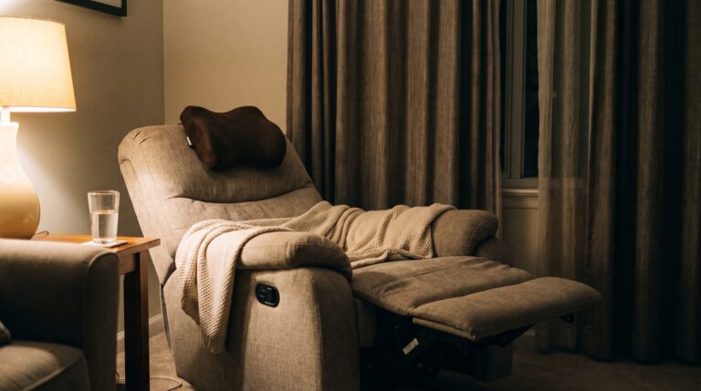 A fabric recliner chair fully reclined with the footrest raised, a throw blanket across the seat and a small contoured neck cushion at the headrest, lit by a warm lamp