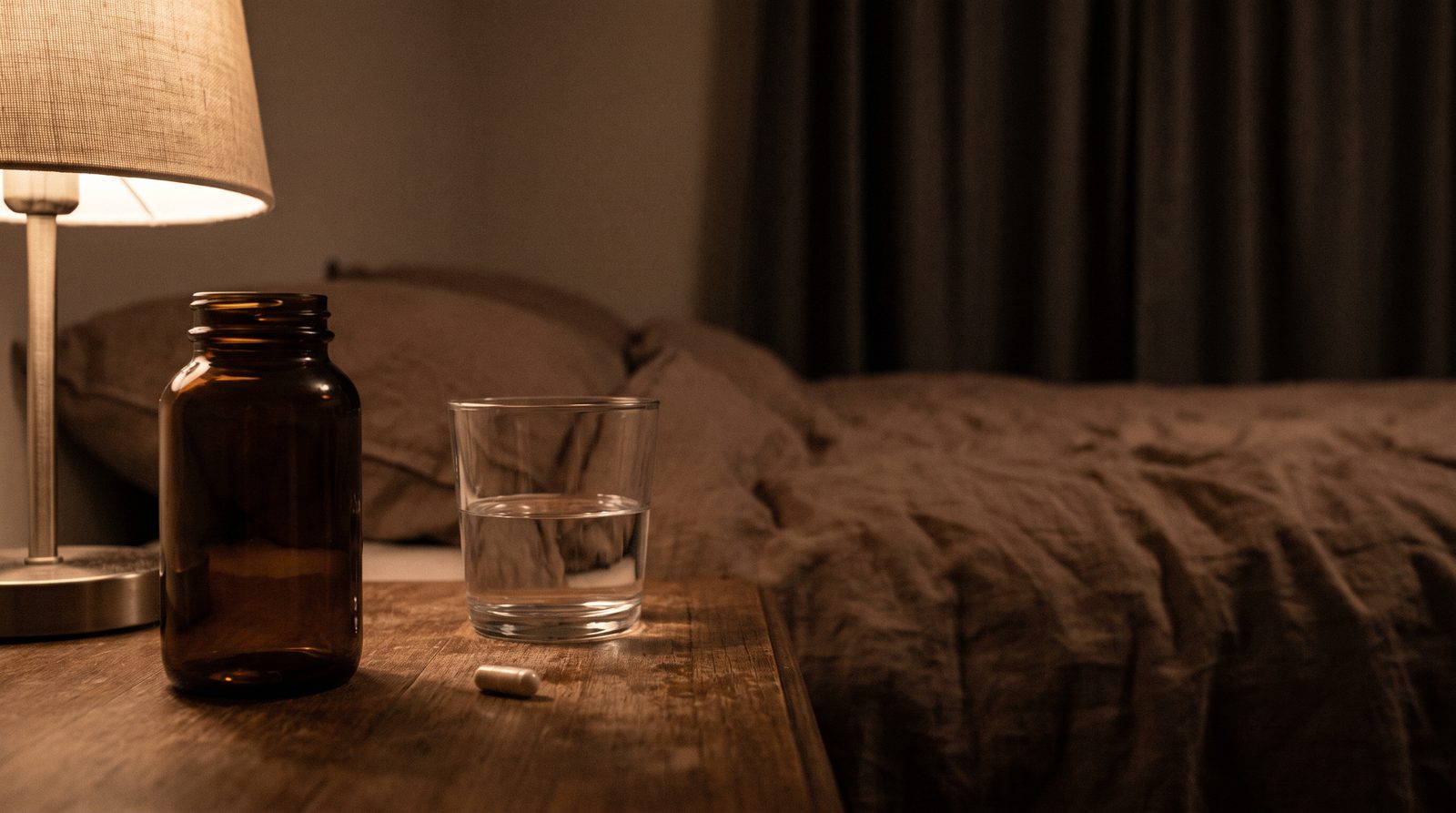 A plain unlabelled amber glass supplement bottle on a bedside table at night beside a glass of water, lit by a low warm lamp