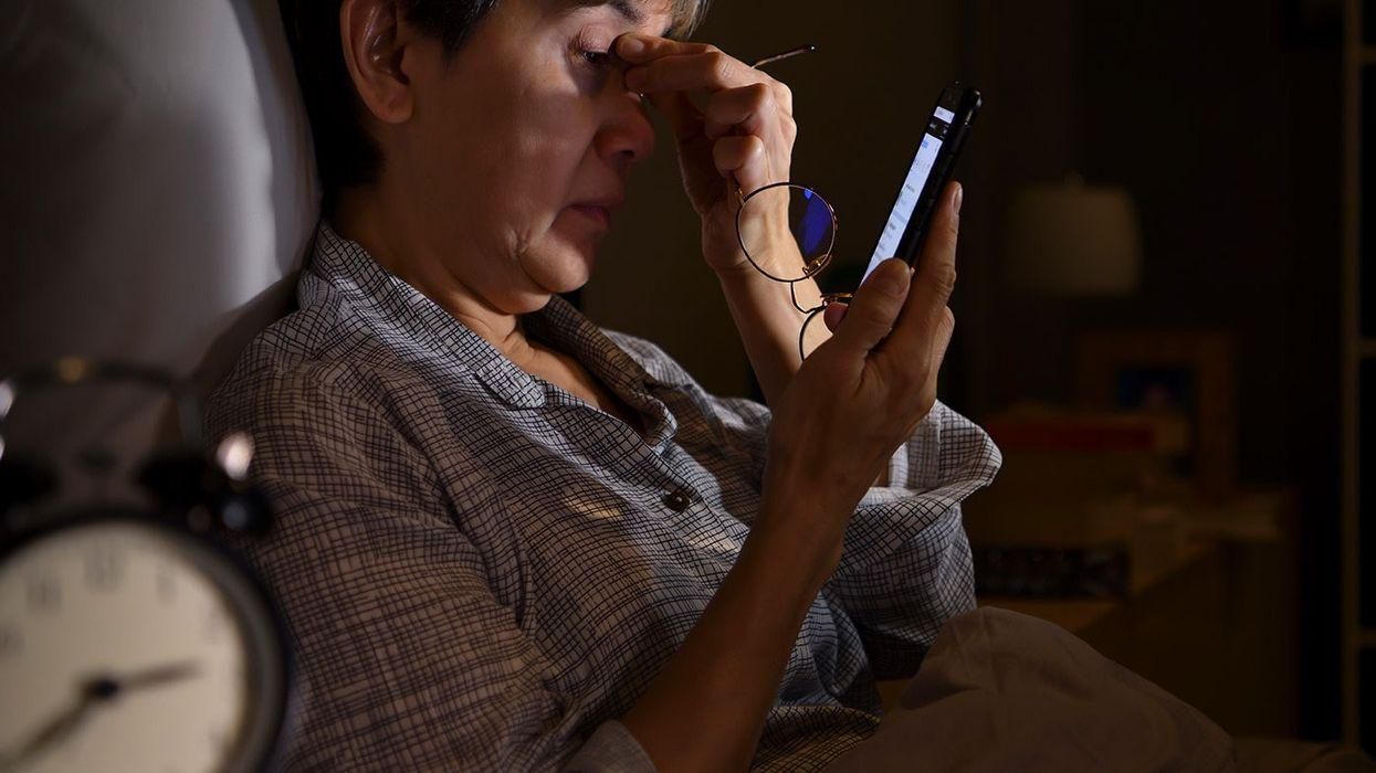Tired woman in bed at night rubbing her eyes while looking at her phone, alarm clock in the foreground