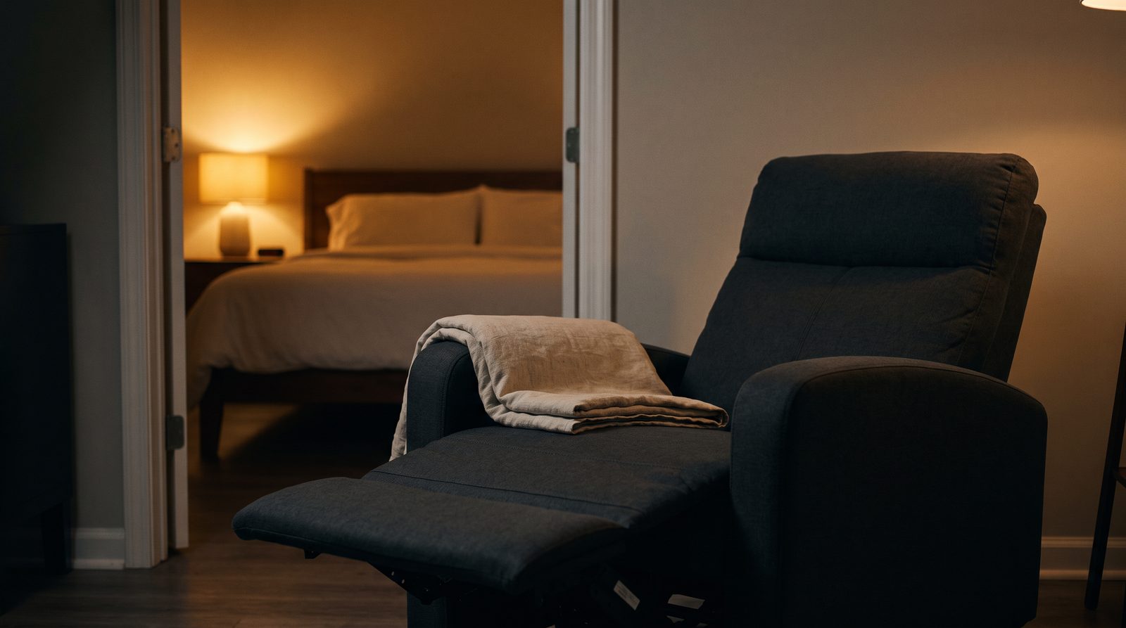 An unbranded fabric recliner with its footrest raised and a folded blanket over the arm, with an empty neatly made bed visible through an open doorway behind it, lit by a warm bedside lamp.