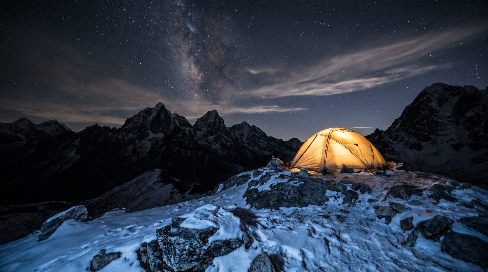 A small mountaineering tent glowing warm amber at night on a high snowy ridge beneath dark peaks and a star-filled sky