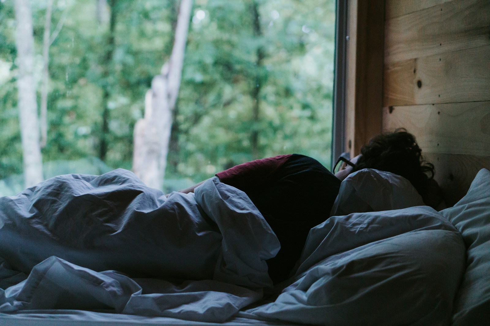 Person lying on their side in a bed with rumpled white bedding beside a window at dusk