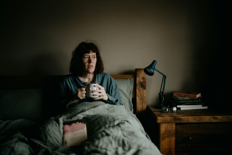 Woman sitting up in bed holding a mug of tea in dim morning light, looking tired