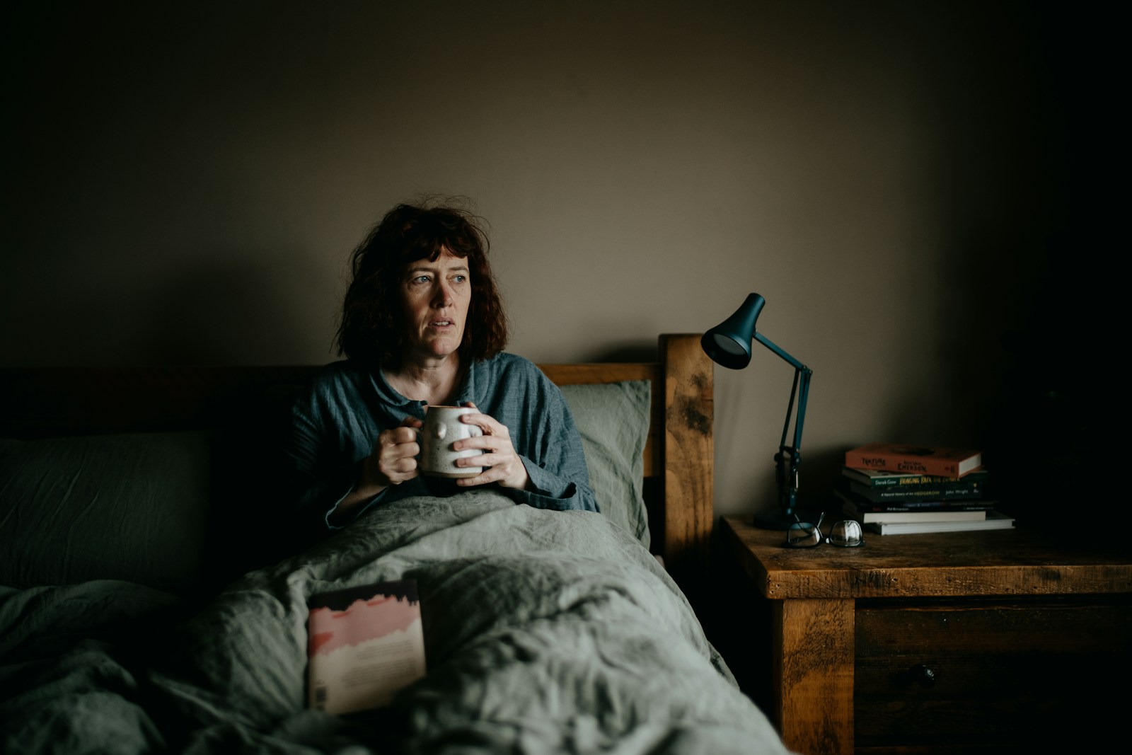 Woman sitting up in bed holding a mug of tea in dim morning light, looking tired