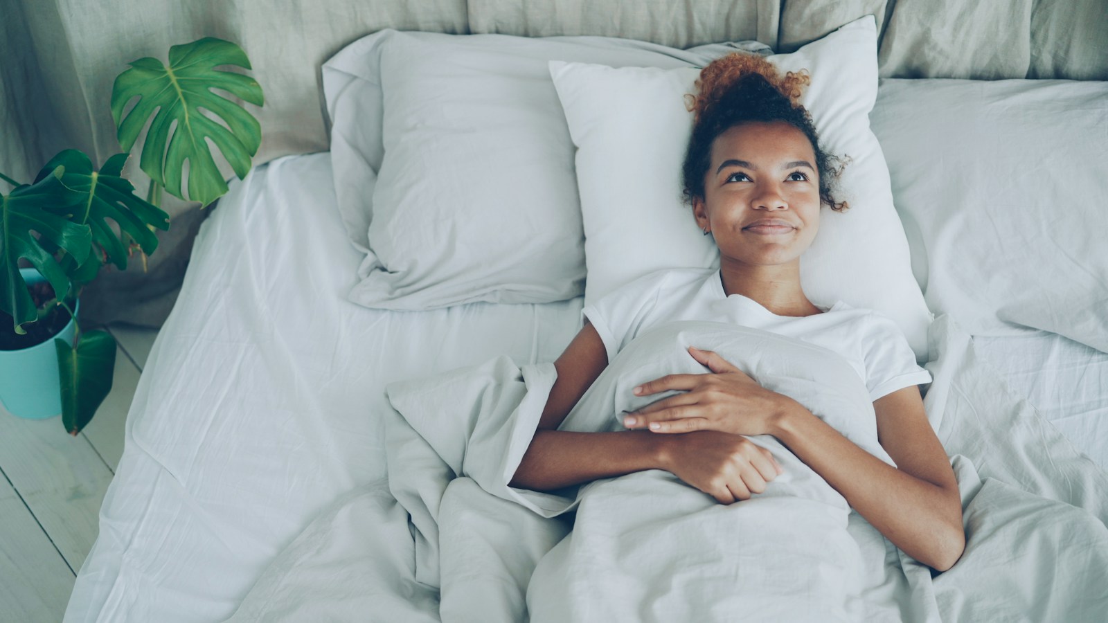 Woman lying in bed under a grey duvet smiling, in a bright bedroom with a plant beside the bed